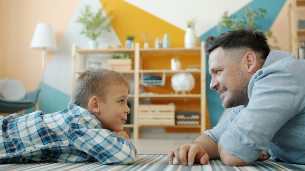 Father and young son lying on the floor at home, talking and smiling together